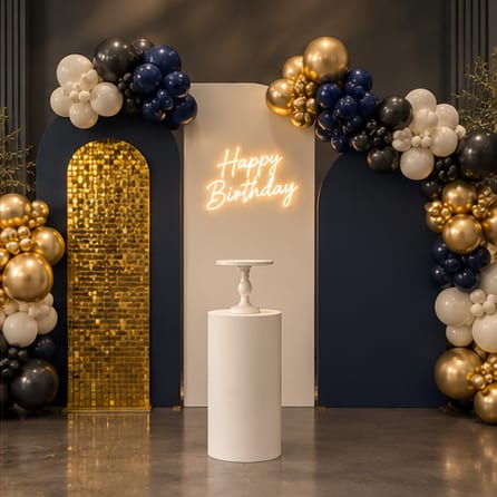 Birthday backdrop of navy, gold-sequin and ivory arched panels with a warm-white neon 'Happy Birthday' sign, a white cylinder plinth with cake stand, and garlands of navy, black, gold and ivory balloons.