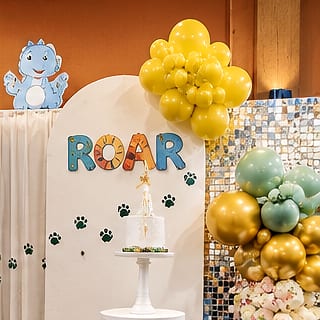 Dinosaur-themed birthday backdrop: white arch with 'ROAR' lettering and paw prints, a cartoon dinosaur cut-out, yellow balloons and a small white cake on a stand.