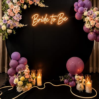 Orange neon 'Bride to Be' sign on a black round backdrop, with purple balloon clusters, pastel flowers and lit pillar candles along a glowing rope light on the floor.