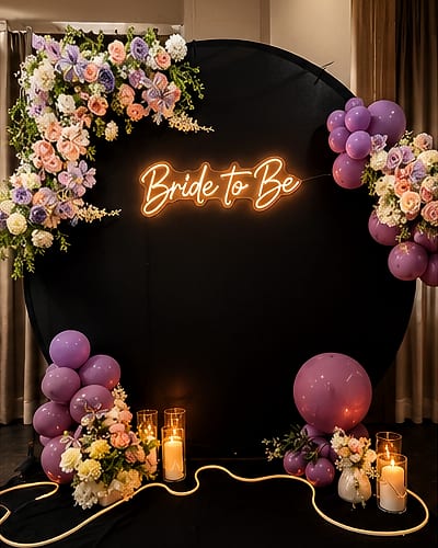 Black round backdrop with an orange neon 'Bride to Be' sign, pastel floral corners, purple balloon clusters and lit pillar candles on the floor with a glowing rope light.